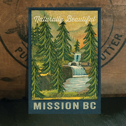 Postcard with a scenic illustration of a forest and waterfall, labeled 'Naturally Beautiful Mission BC', on a wooden surface.